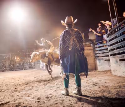 Bull rider on a bucking bull at night with bullfighter and crowd at a rodeo arena