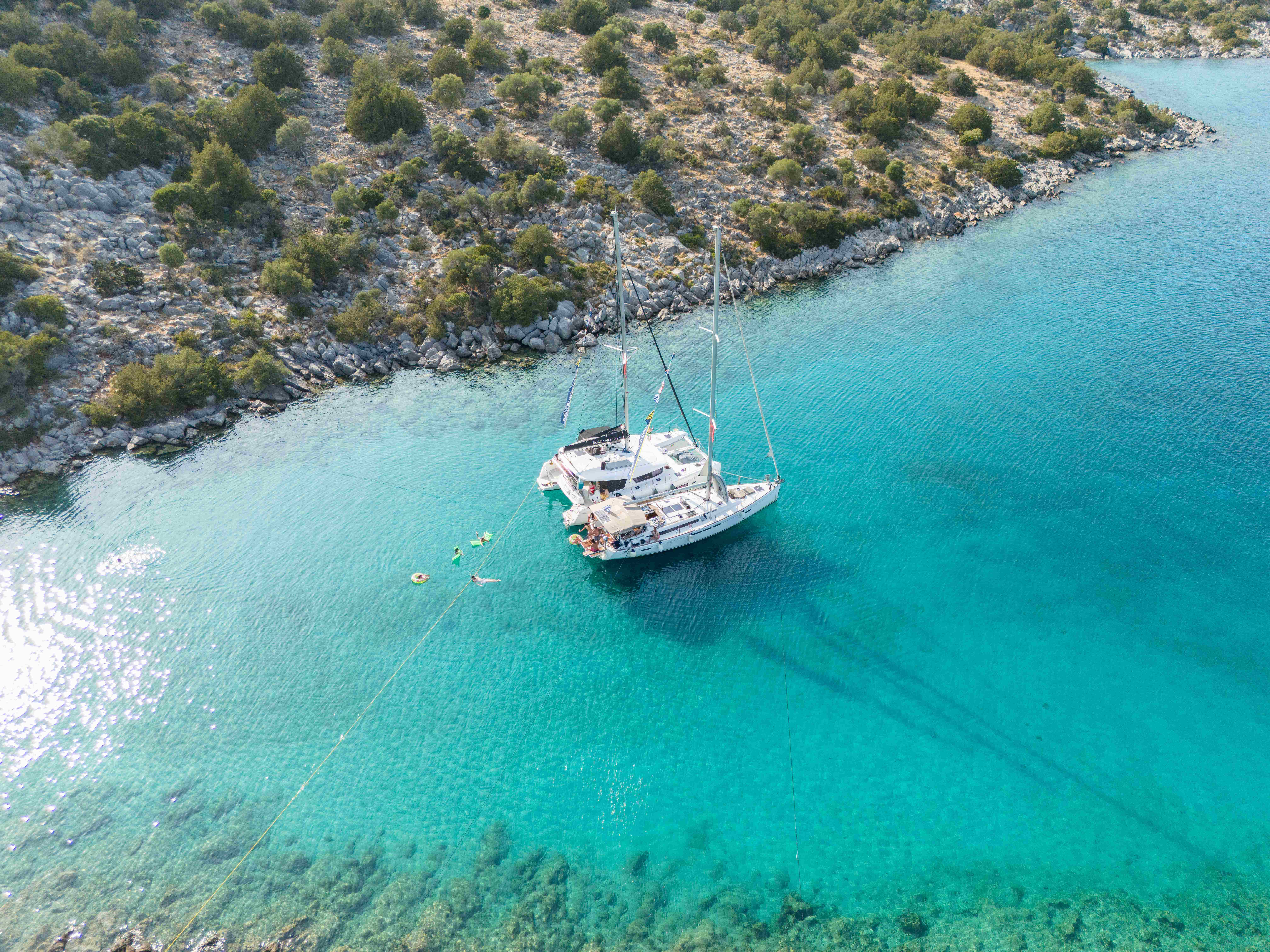 Aerial side view of a catamaran and sailboat with people relaxing in turquoise waters in Greece