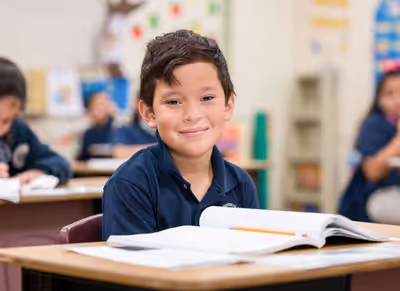 Smiling elementary school boy in navy uniform sits at desk with open workbook and pencil in classroom