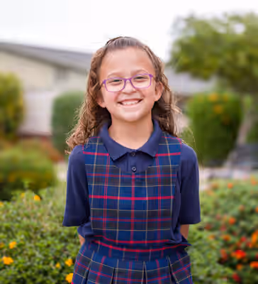 Smiling elementary school girl wearing plaid school uniform and glasses stands outside on a private school campus with green landscaping in the background