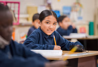Smiling elementary school girl in navy blue uniform writes in a workbook at her desk during class