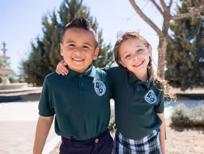 Two elementary school students in matching green uniforms smile with arms around each other on a sunny day outside their school campus, standing in front of trees and a brick path