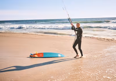 Kitesurfer holding control bar at water's edge with kiteboard on wet sand
