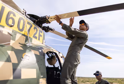 Pilot performing pre-flight rotor blade inspection on camouflaged MD 530G helicopter