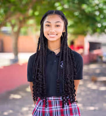 Teenage girl in black polo and plaid skirt smiles confidently on campus